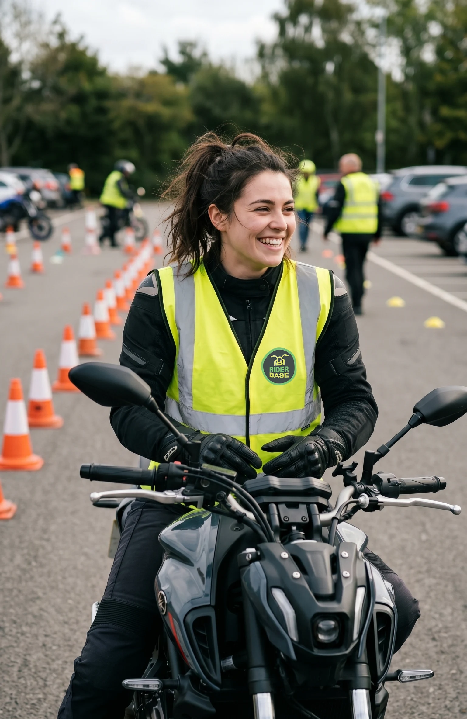 A smiling young woman in black motorcycle gear and a yellow high-visibility vest sits on a dark grey motorcycle.